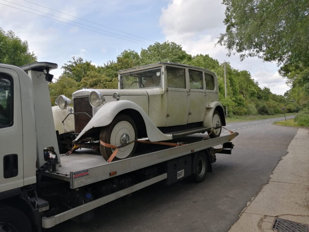 An old car is being towed on a flatbed truck, showcasing its vintage design against a clear blue sky.
